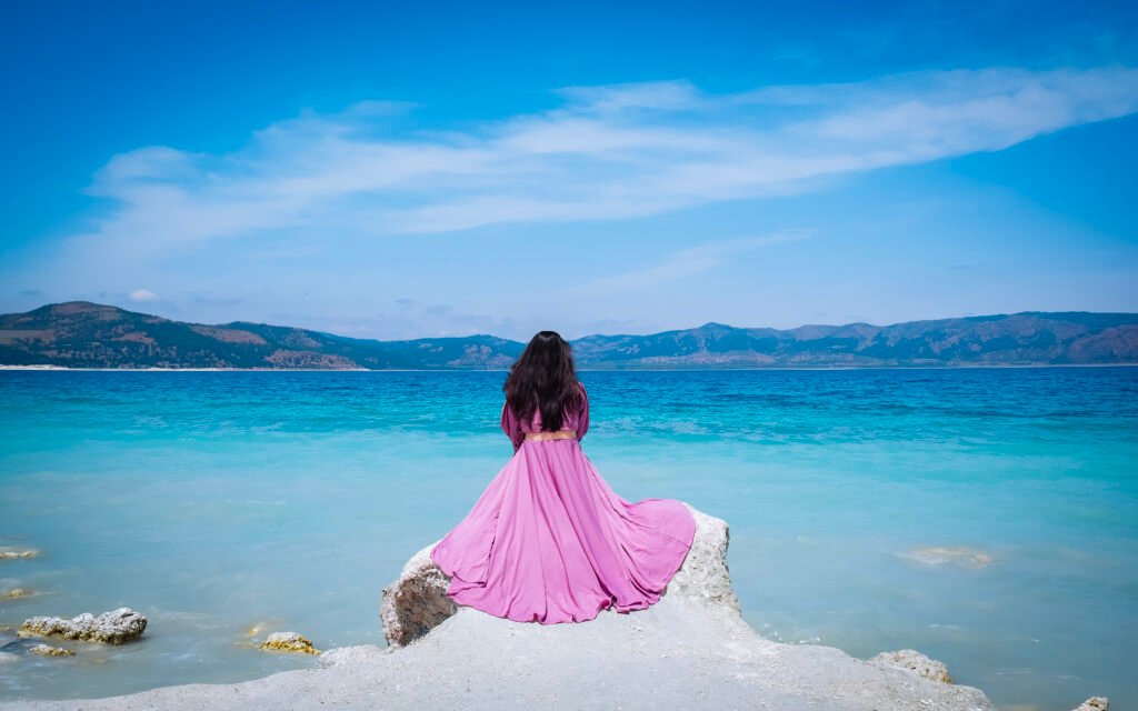 Image of a woman in a pink gown sitting on the bank of a blue lake.