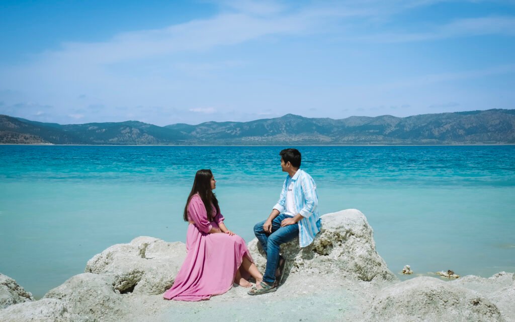 An image of a couple sitting on a white rock beside a blue lake.