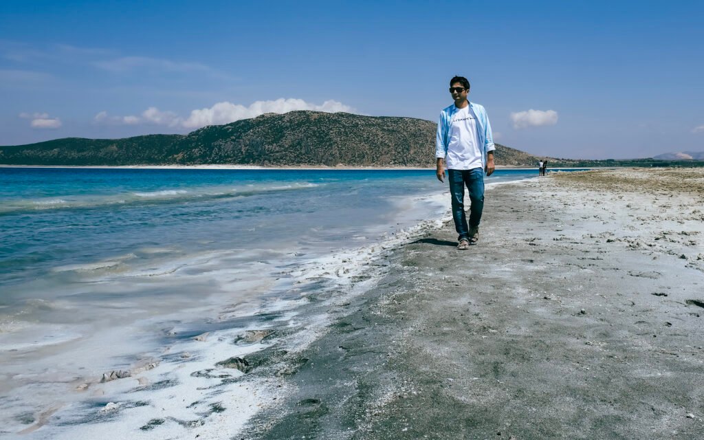 Image of a man walking along the shore of a crystal-clear blue lake with black sand.